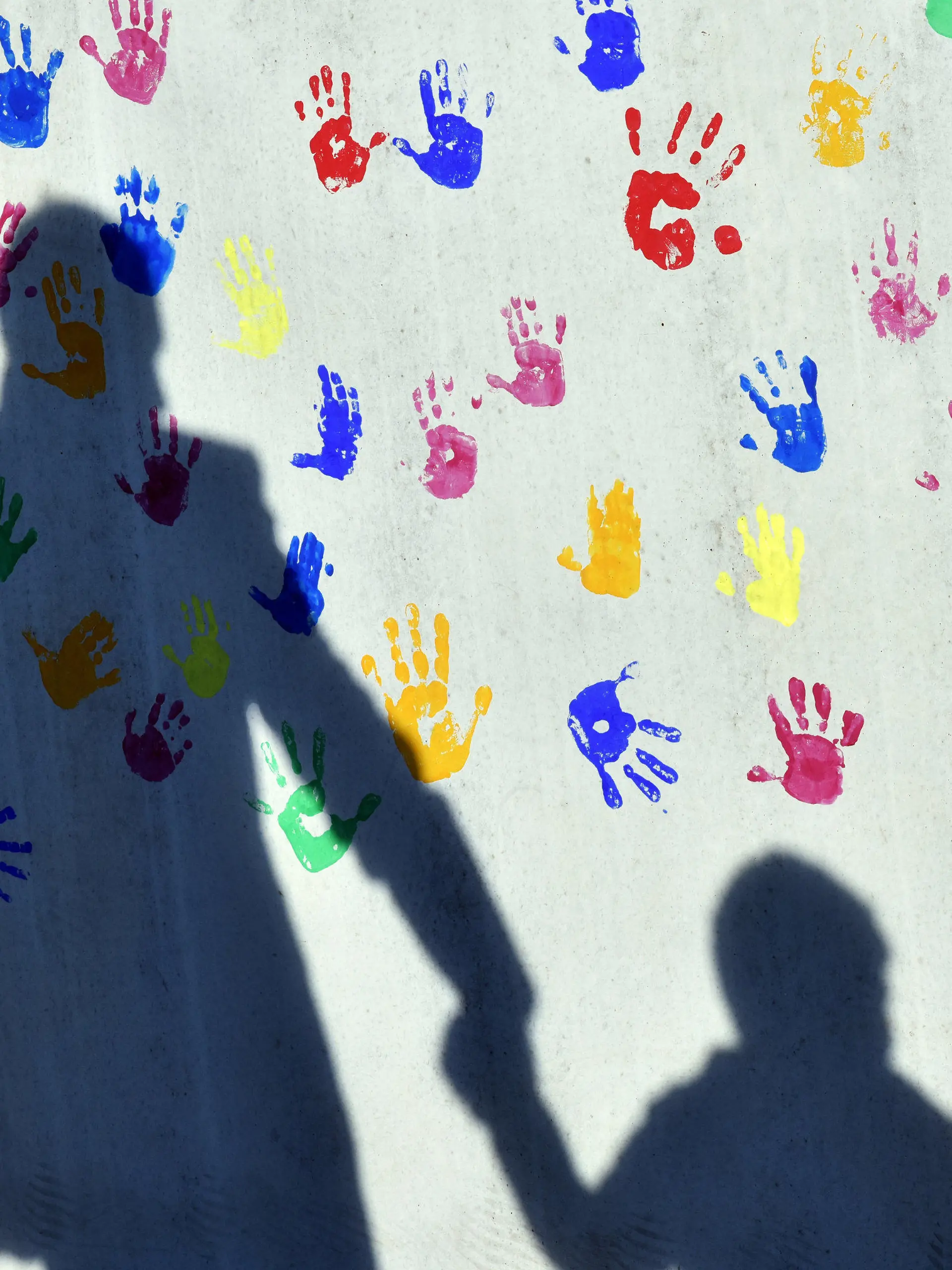 ARCHIV - 08.04.2015, Bayern, München: Ein Mann mit einem Kind auf dem Arm und einem an der Hand wirft einen Schatten auf eine mit bunten Handabdrücken bemalte Wand einer Kindertagesstätte. (zu dpa «Der Corona-Doppeljob trifft Kita-Eltern hart - und Mütter besonders?») Foto: Peter Kneffel/dpa +++ dpa-Bildfunk +++