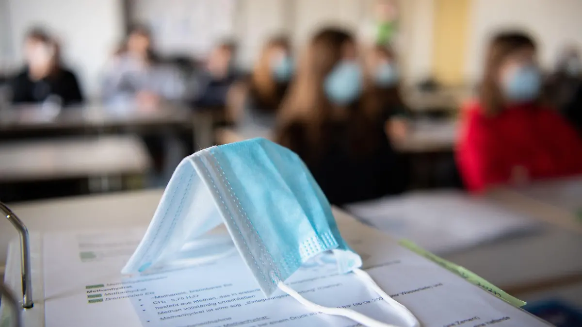 ARCHIV - 17.11.2020, Bayern, München: Eine Maske liegt im Unterricht auf Unterlagen, während im Hintergrund Schülerinnen und Schüler in einem Gymnasium mit Mund- und Nasenschutz dem Unterricht folgen. Die Corona-Inzidenzen bei Kindern und Jugendlichen in Bayern sind so hoch wie nie zuvor.       (zu dpa «Rekord-Inzidenz bei bayerischen Kindern und Jugendlichen») Foto: Matthias Balk/dpa +++ dpa-Bildfunk +++
