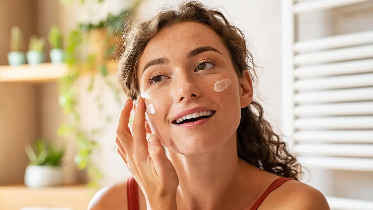 Woman caring of her beautiful skin face standing near mirror in the bathroom. Young woman applying moisturizing cream on her face during morning routine. Smiling natural girl holding little green jar of ecological cosmetic cream.
