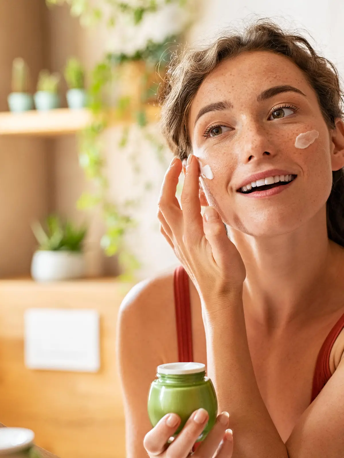 Woman caring of her beautiful skin face standing near mirror in the bathroom. Young woman applying moisturizing cream on her face during morning routine. Smiling natural girl holding little green jar of ecological cosmetic cream.