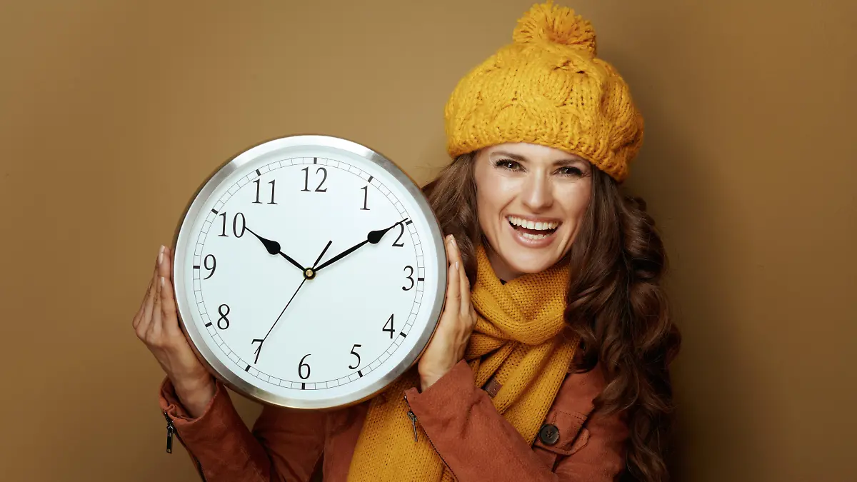 happy elegant woman in yellow beret and scarf showing round clock remind about autumn time change against brown background.