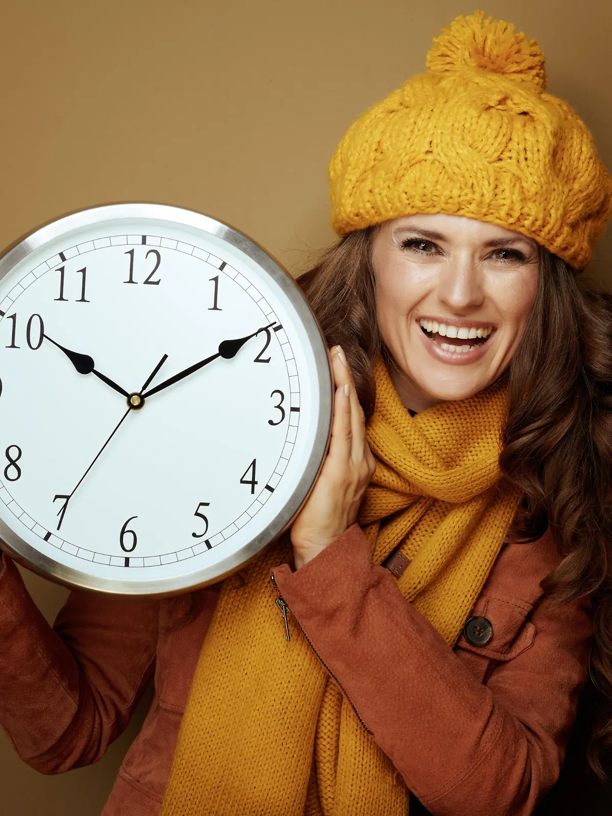 happy elegant woman in yellow beret and scarf showing round clock remind about autumn time change against brown background.