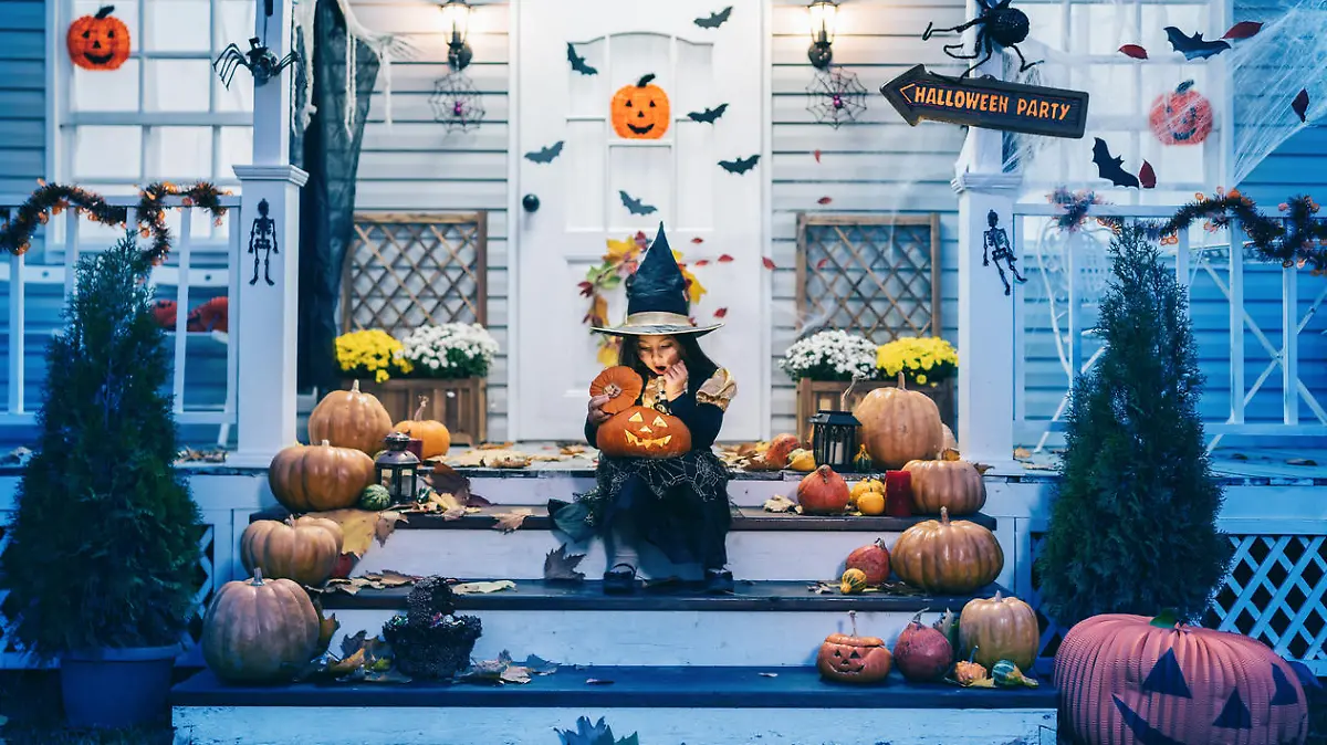 Little girl in witch costume sitting on the stairs in front of the house and holding Jack-o-Lantern Pumpkins on Halloween trick or treat