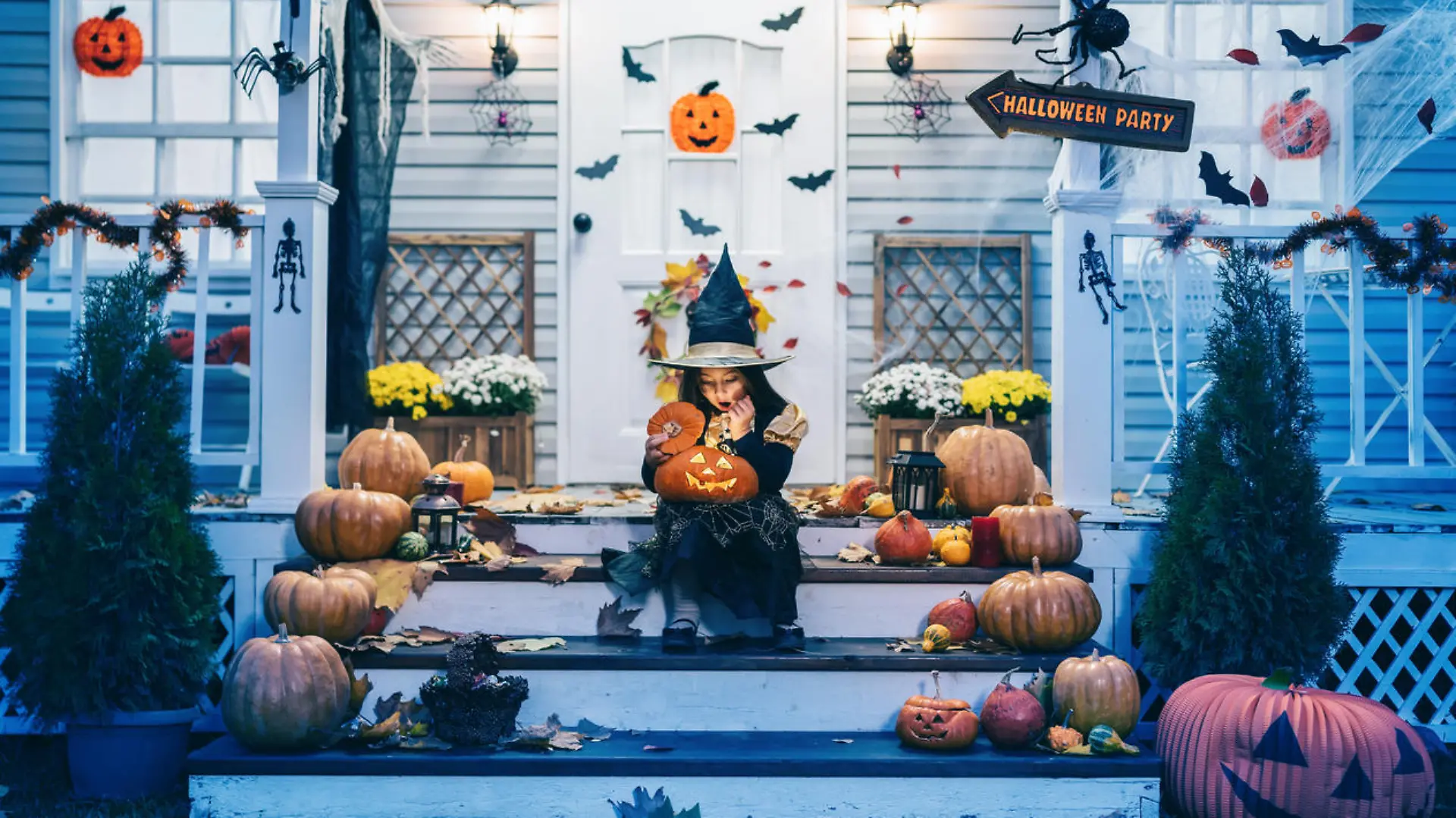 Little girl in witch costume sitting on the stairs in front of the house and holding Jack-o-Lantern Pumpkins on Halloween trick or treat