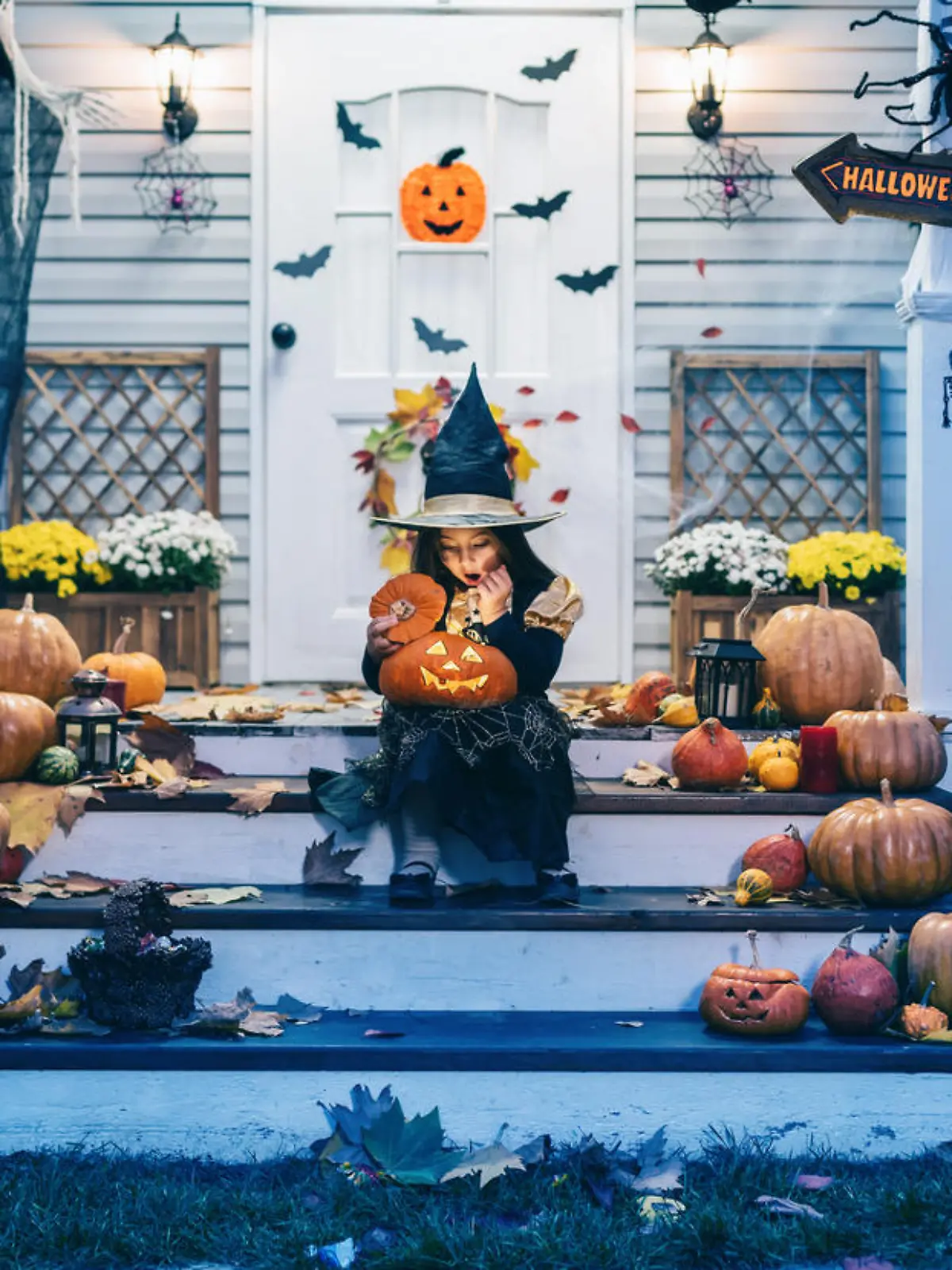 Little girl in witch costume sitting on the stairs in front of the house and holding Jack-o-Lantern Pumpkins on Halloween trick or treat