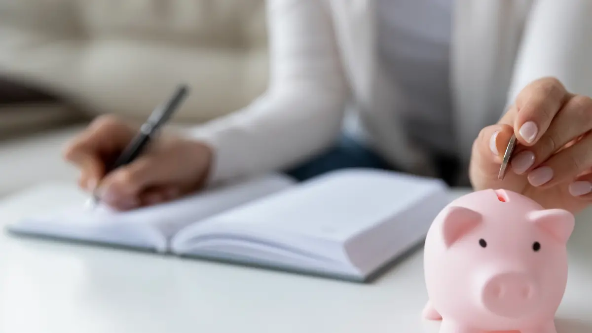 Thrifty woman sit at table hold pen writing daily expenses in diary put coin in pink piggy bank close up. Saving money for future, caring for tomorrow, makes stash of cash, investment, economy concept