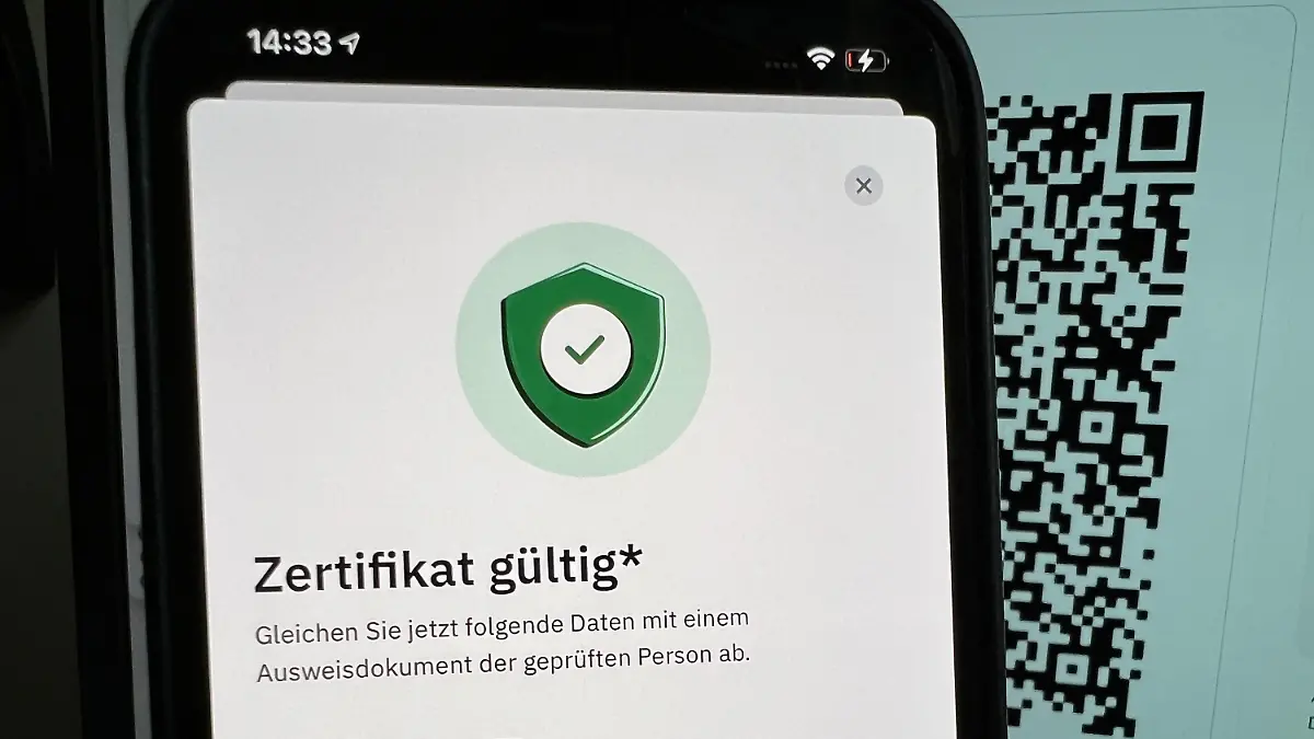 27.10.2021, Berlin: ILLUSTRATION - Die App «CovPass Check» zur Überprüfung von digitalen Impfnachweisen weist ein gefälschtes Zertifikat für «Adolf Hitler» auf einem Smartphone als echt aus, wobei im Hintergrund der QR-Code des gefälschten Impfzertifikats auf einem Computerbildschirm zu sehen ist (gestellte Aufnahme). Im Internet war das gefälschte digitale Impfzertifikat aufgetaucht. Unklar blieb am Mittwoch, ob für das Fake-Zertifikat geheim gehaltene Digitalschlüssel einer Ausgabestelle entwendet wurden. (Zu dpa «Fake-Impfzertifikat für «Adolf Hitler» als echt ausgewiesen») Foto: Christoph Dernbach/dpa +++ dpa-Bildfunk +++
