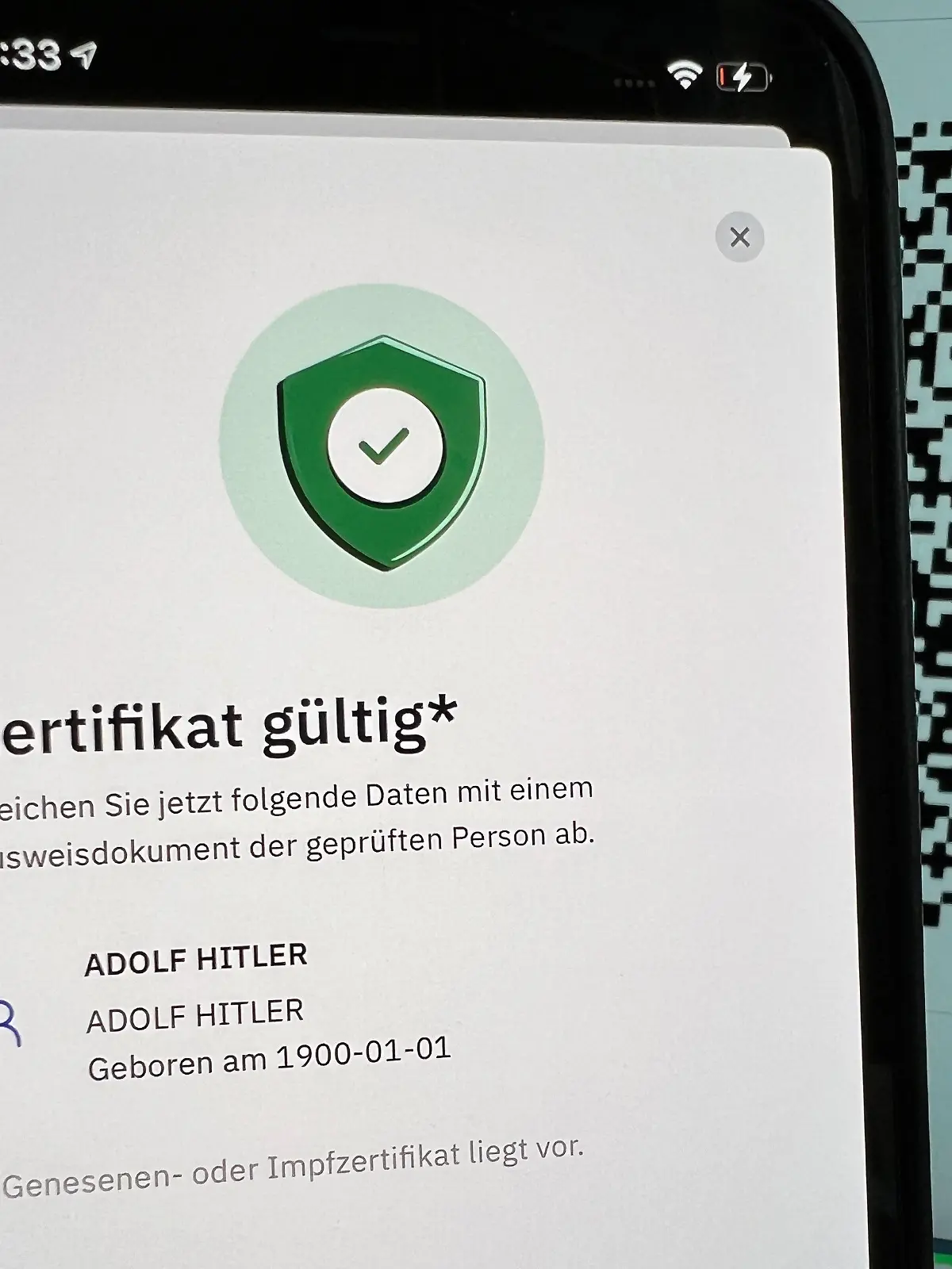 27.10.2021, Berlin: ILLUSTRATION - Die App «CovPass Check» zur Überprüfung von digitalen Impfnachweisen weist ein gefälschtes Zertifikat für «Adolf Hitler» auf einem Smartphone als echt aus, wobei im Hintergrund der QR-Code des gefälschten Impfzertifikats auf einem Computerbildschirm zu sehen ist (gestellte Aufnahme). Im Internet war das gefälschte digitale Impfzertifikat aufgetaucht. Unklar blieb am Mittwoch, ob für das Fake-Zertifikat geheim gehaltene Digitalschlüssel einer Ausgabestelle entwendet wurden. (Zu dpa «Fake-Impfzertifikat für «Adolf Hitler» als echt ausgewiesen») Foto: Christoph Dernbach/dpa +++ dpa-Bildfunk +++