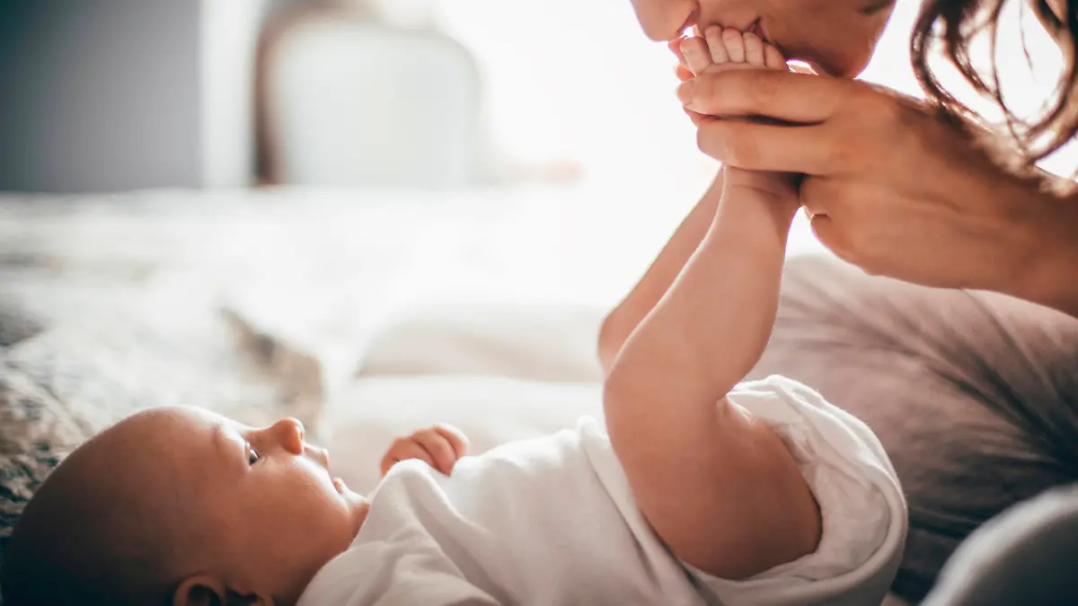 A happy mother kisses her baby's feet while she is lying down. The baby is looking up at her wearing a white baby grow.
