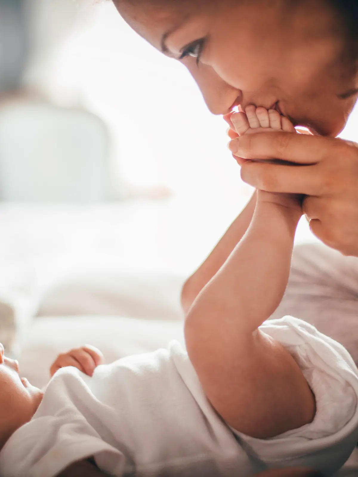 A happy mother kisses her baby's feet while she is lying down. The baby is looking up at her wearing a white baby grow.