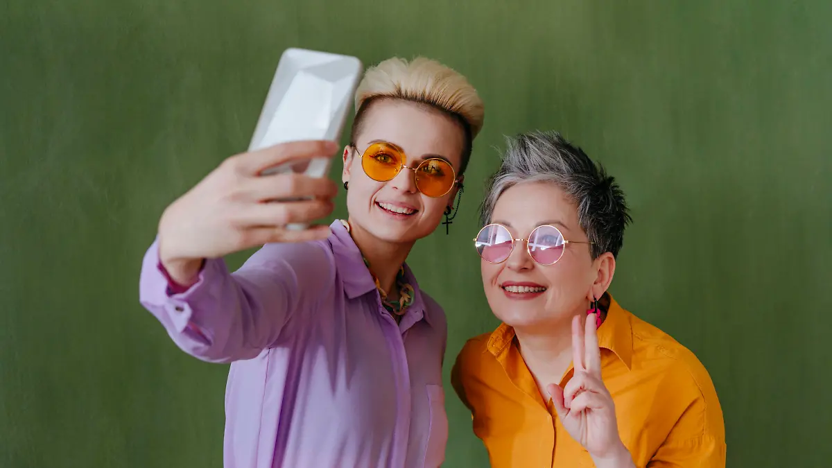 Happy women taking selfie through smart phone in front of green wall || Modellfreigabe vorhanden