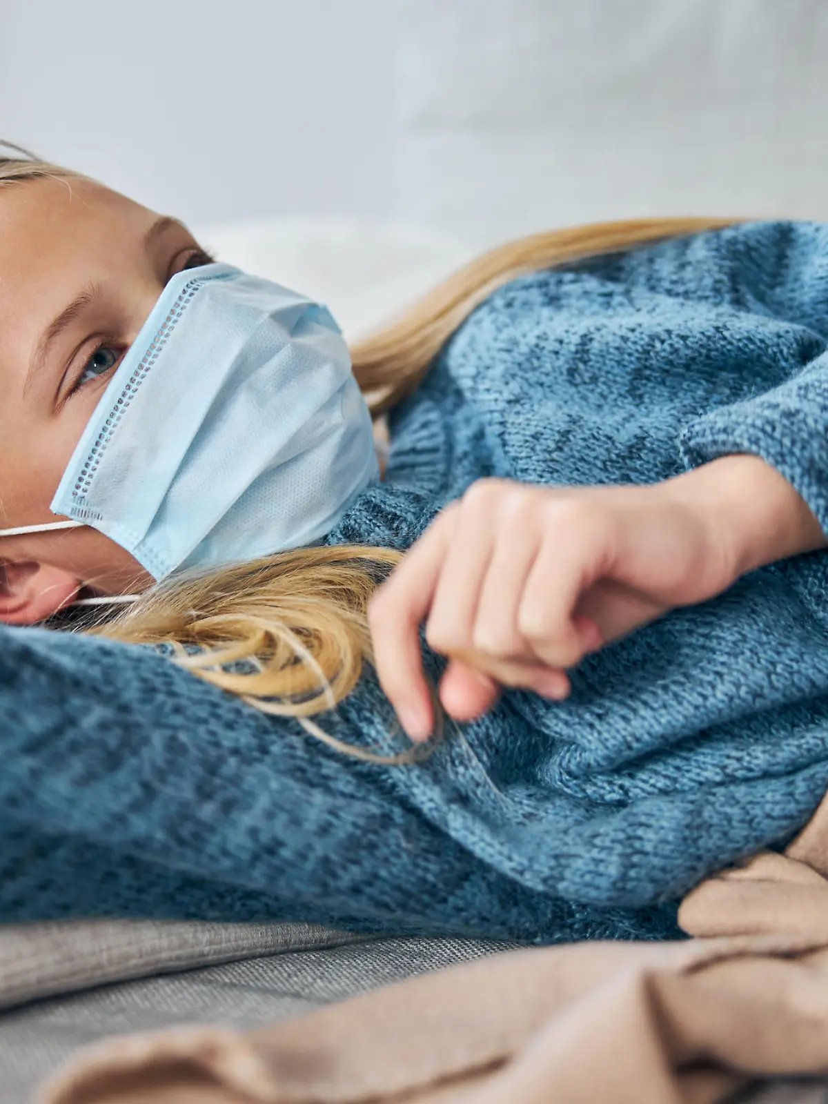 Low-spirited female child in a face mask lying on the sofa at home in broad daylight