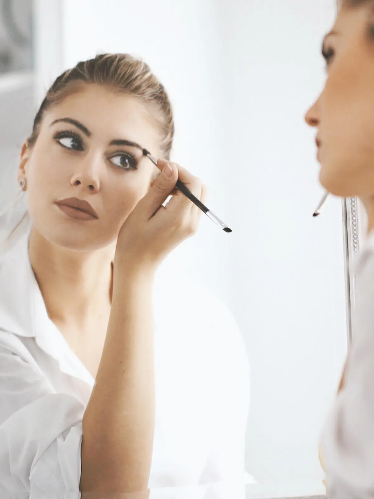 Closeup rear view of attractive mid 20's blond woman putting on some makeup before heading to work. She's in front of large bathroom mirror, focus is on her reflection.