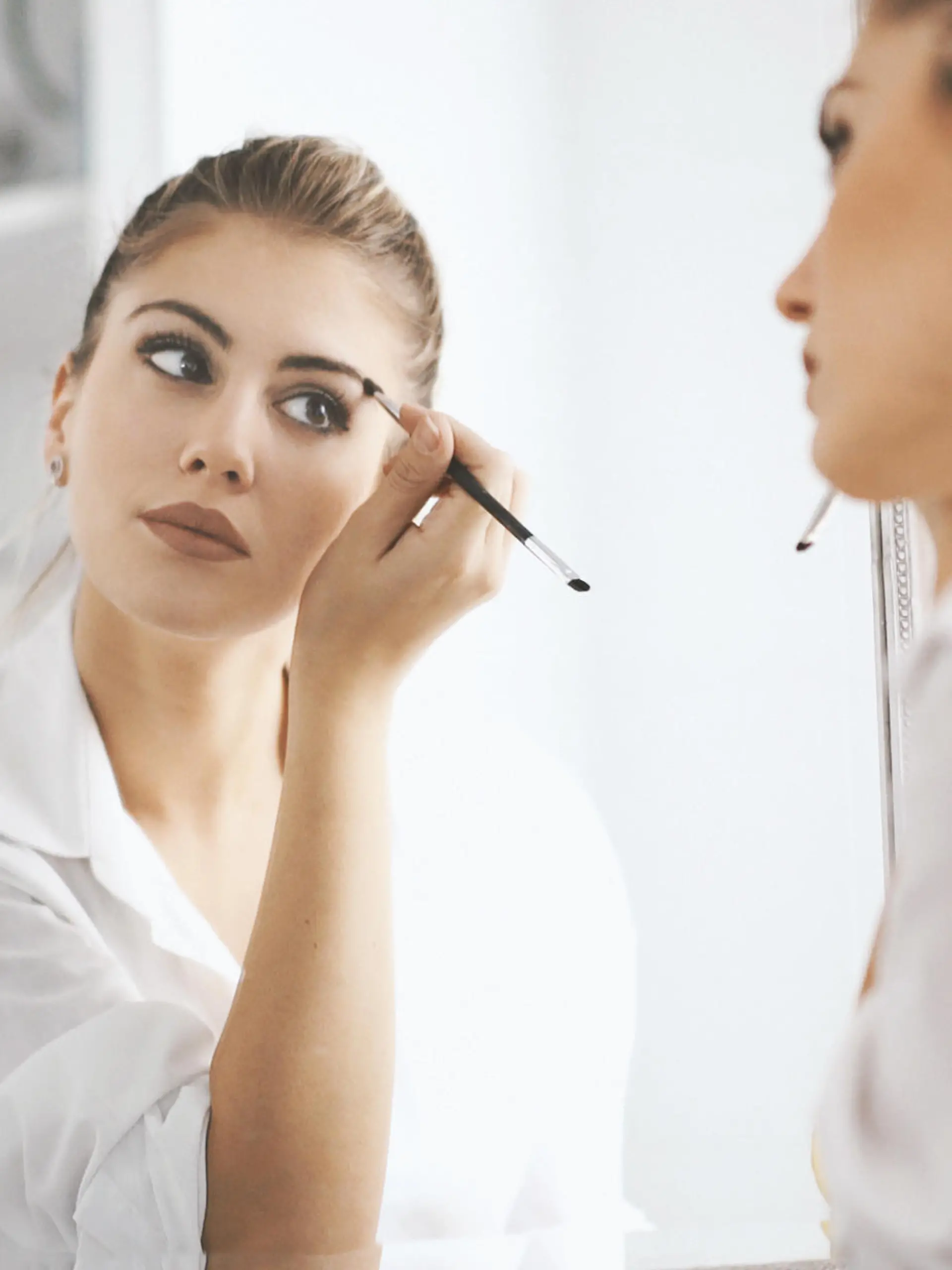 Closeup rear view of attractive mid 20's blond woman putting on some makeup before heading to work. She's in front of large bathroom mirror, focus is on her reflection.