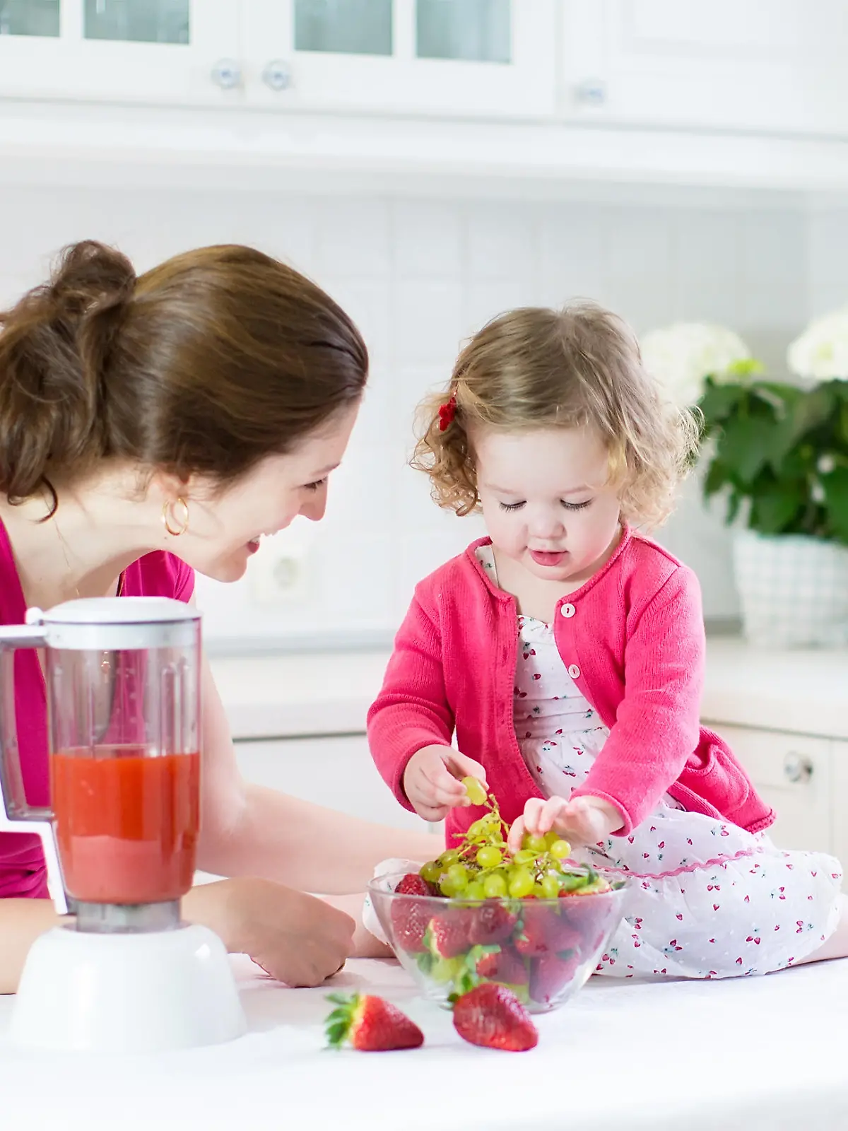 Beautiful young mother and her happy laughing toddler girl making fresh strawberry and other fruit juice for breakfast together in a sunny white kitchen with a window