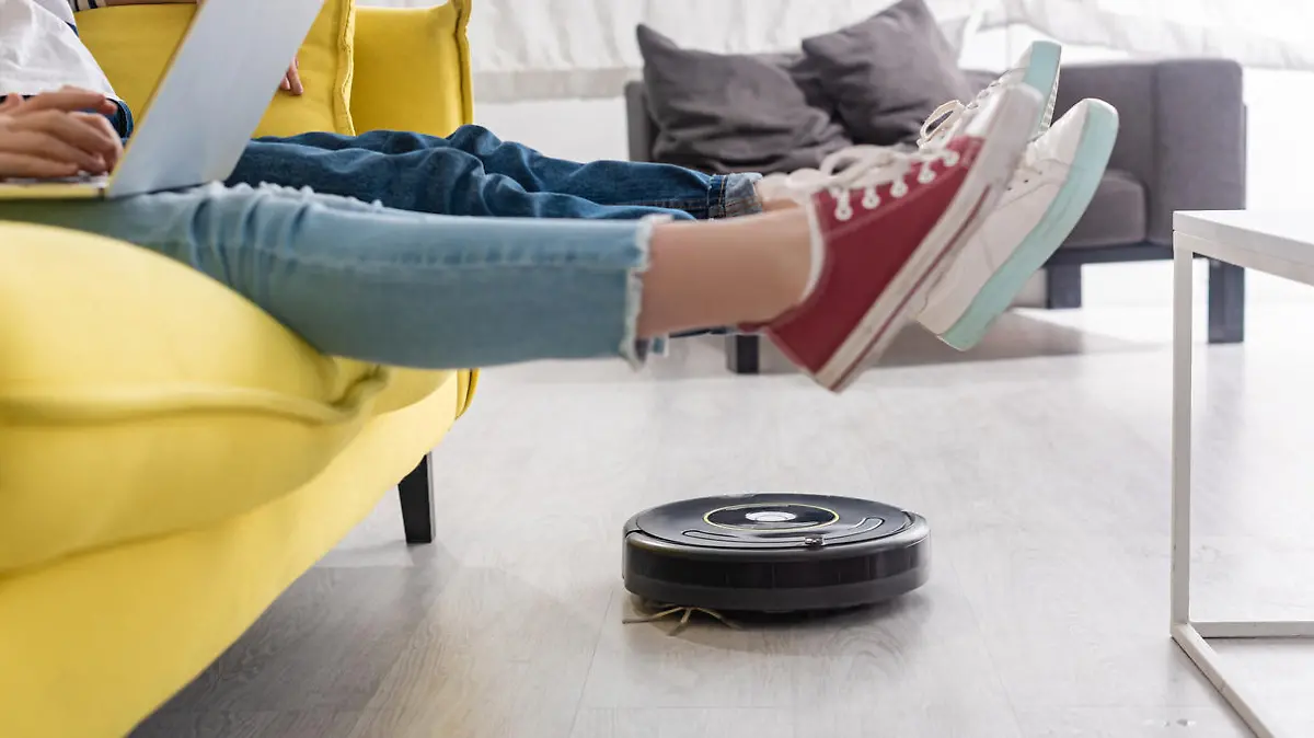 Cropped view of kid with laptop near mother with raised legs on sofa and robotic vacuum cleaner on floor in living room