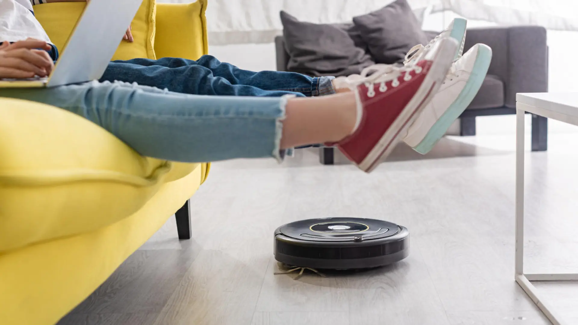 Cropped view of kid with laptop near mother with raised legs on sofa and robotic vacuum cleaner on floor in living room