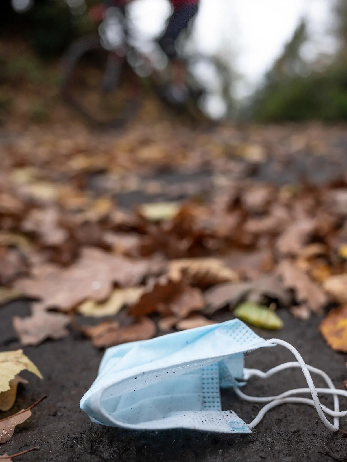 ARCHIV - 01.11.2020, Nordrhein-Westfalen, Herdecke: Eine weggeworfene Maske liegt zwischen Herbstlaub auf einer Straße, während ein Radfahrer vorbeifährt. Nordrhein-Westfalen lockert weiter. Im Freien müssen keine Masken mehr getragen werden. Und auch bei  Veranstaltungen und in der Gastronomie gibt es weitere Lockerungen. (zu dpa: «Keine Maskenpflicht mehr im Freien») Foto: Bernd Thissen/dpa +++ dpa-Bildfunk +++
