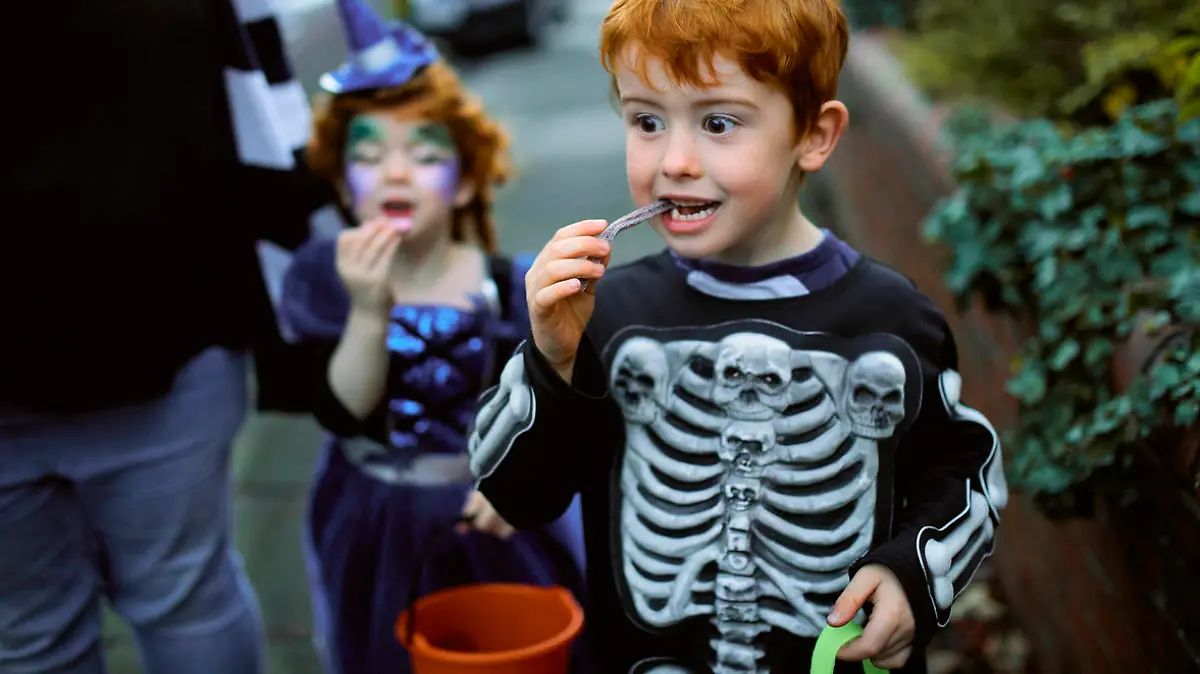 Little boy eating halloween candy while trick or treating in suburban streets with his mother and sister.