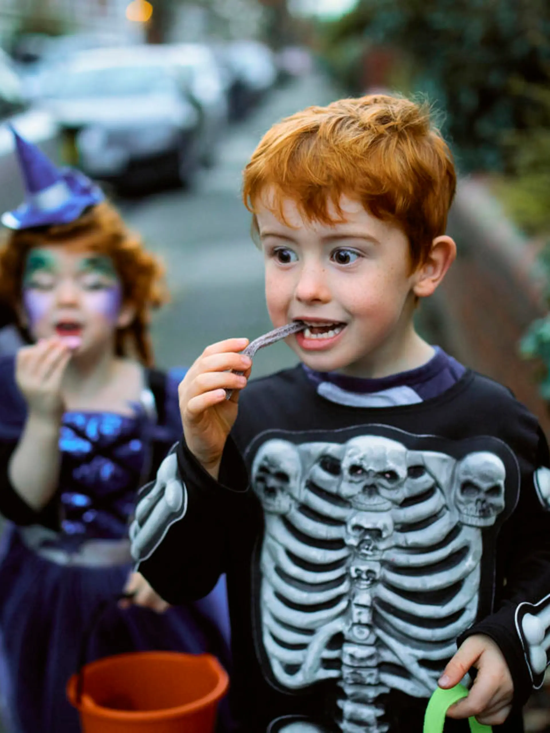 Little boy eating halloween candy while trick or treating in suburban streets with his mother and sister.