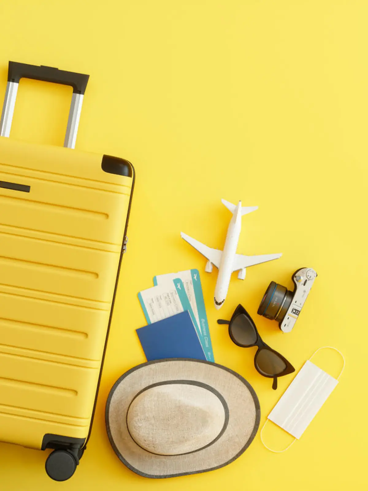 Medical Mask, Suitcase with Sun Hat, Camera, Passport, Airplane Ticket, Sunglasses and Airplane on Yellow Background