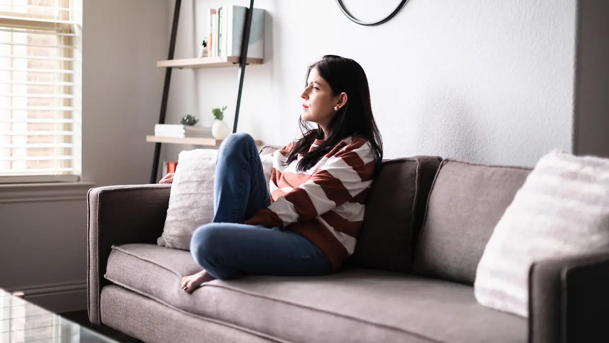 A sad mid adult Caucasian woman looks through the window while sitting on a sofa in her home.