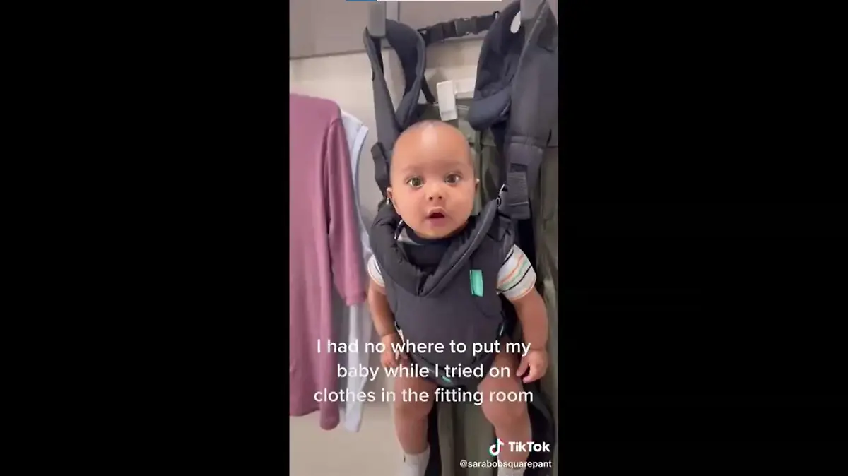 Ein Baby hängt in seiner Trage an der Garderobe einer Umkleidekabine.