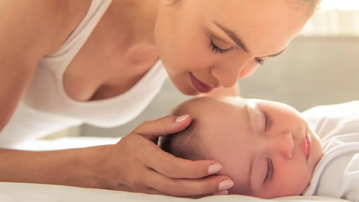 Beautiful young mom is kissing her cute little baby who is sleeping in bed at home, close-up