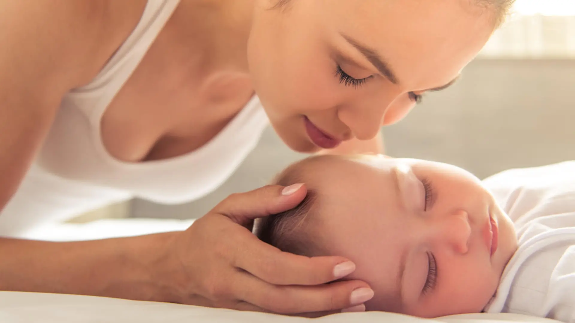 Beautiful young mom is kissing her cute little baby who is sleeping in bed at home, close-up
