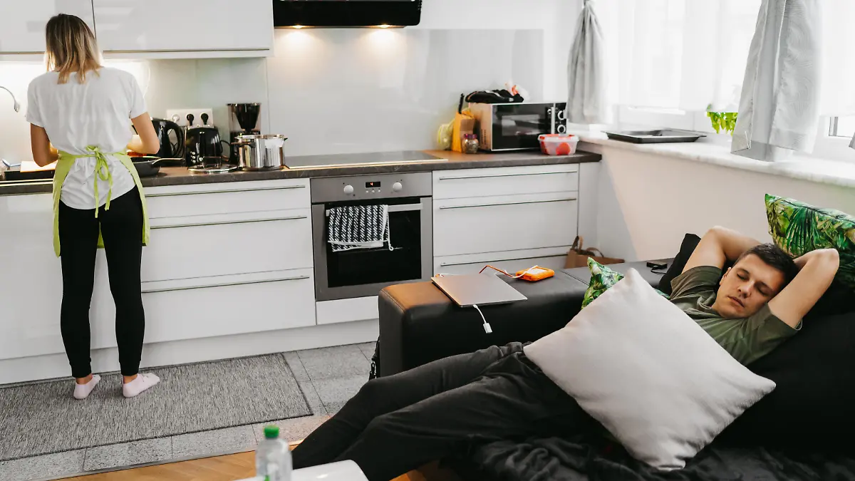Young woman preparing food in kitchen while man sleeping on sofa