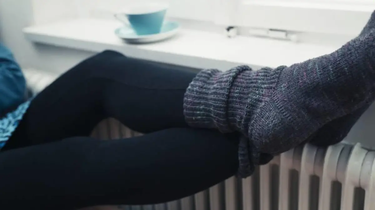 Frau mit dicken Socken und Tasse Tee legt Beine auf Heizung