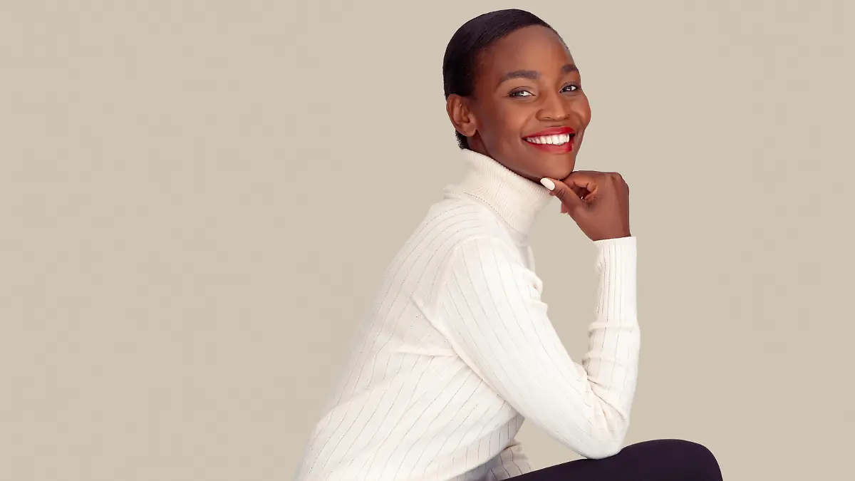 Beautiful black woman wearing white sweater isolated on creamy background. Mid adult stylish woman sitting against light gray wall. Mature fashionable lady with turtle neck sweater smiling with hand on chin while looking at camera with copy space.
