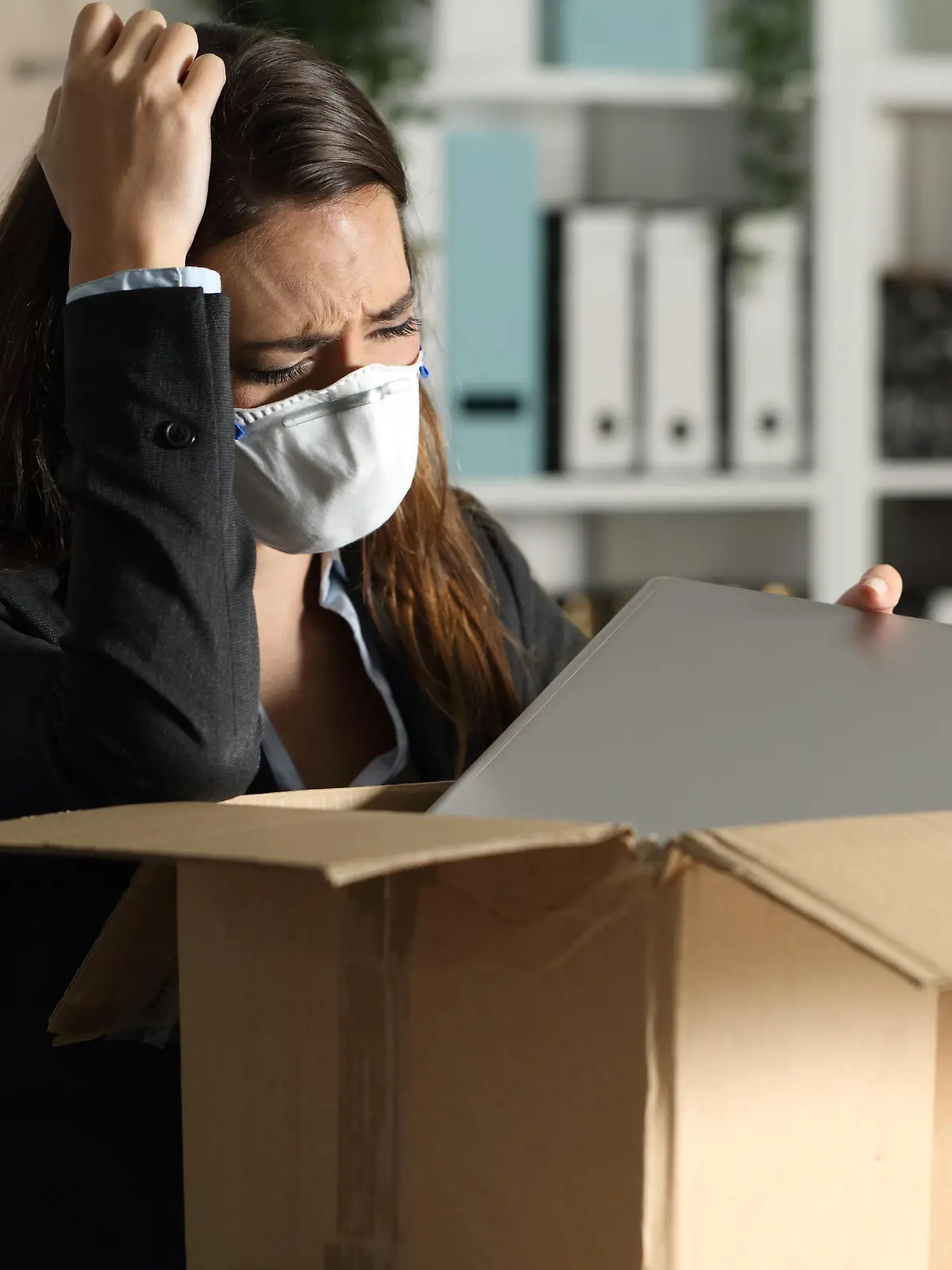 Fired executive woman with protective mask packing personal belongings on a box at the office at night