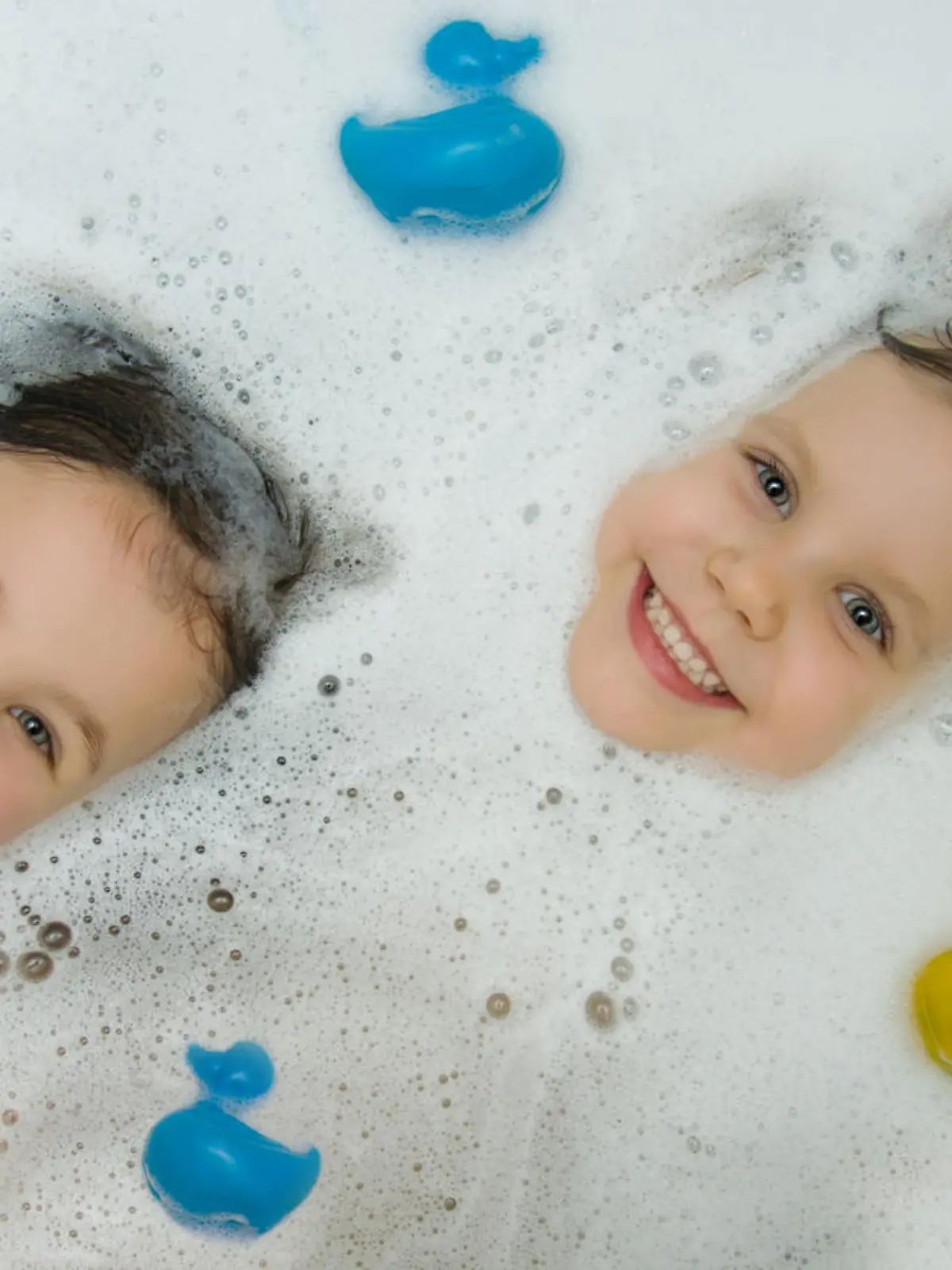 Two cute girls bathe together in a bath with ducks for bathing. The playful faces of children peek out of the foam, smiling cheerfully looking at the camera. View from above. Children's hygiene.