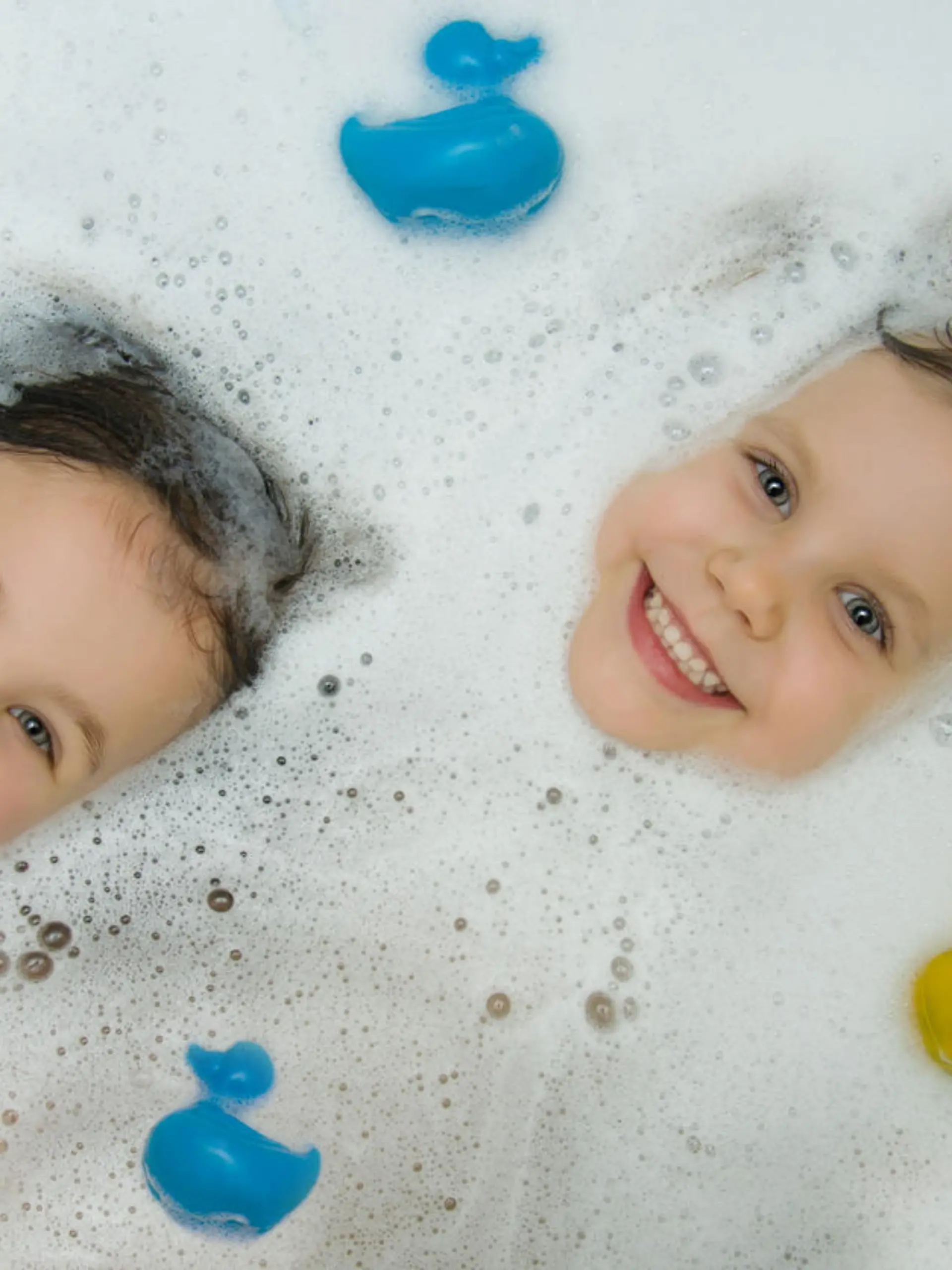 Two cute girls bathe together in a bath with ducks for bathing. The playful faces of children peek out of the foam, smiling cheerfully looking at the camera. View from above. Children's hygiene.