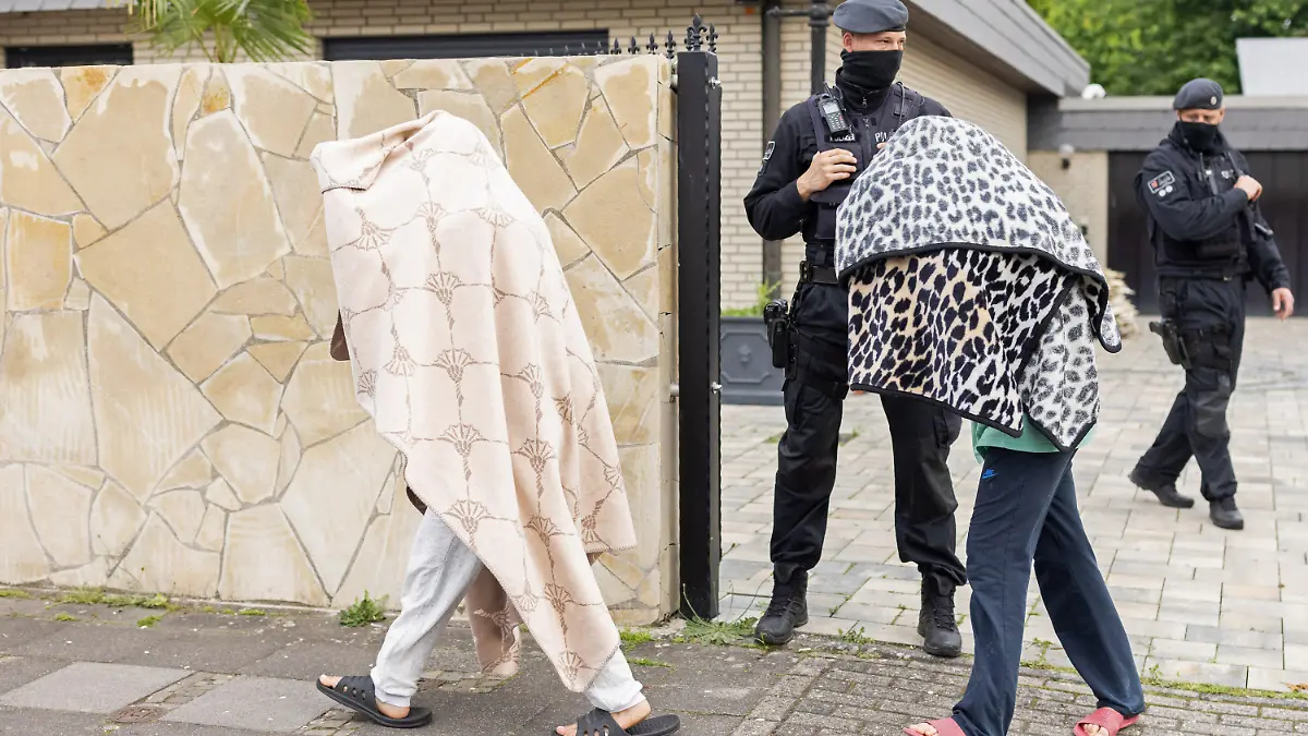 08.06.2021, Nordrhein-Westfalen, Leverkusen: Polizisten stehen vor einer Villa, während zwei Personen mit Decken über dem Oberkörper das Gelände verlassen. Bei Ermittlungen gegen Clankriminalität durchsuchten Spezialkräfte der Polizei rund 30 Objekte