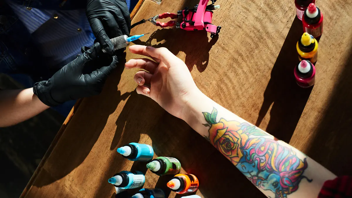 Tattoo expert in gloves holding tattoo gun while hand of female client with new colorful tattoo lying in wooden table beside, high angle close-up view