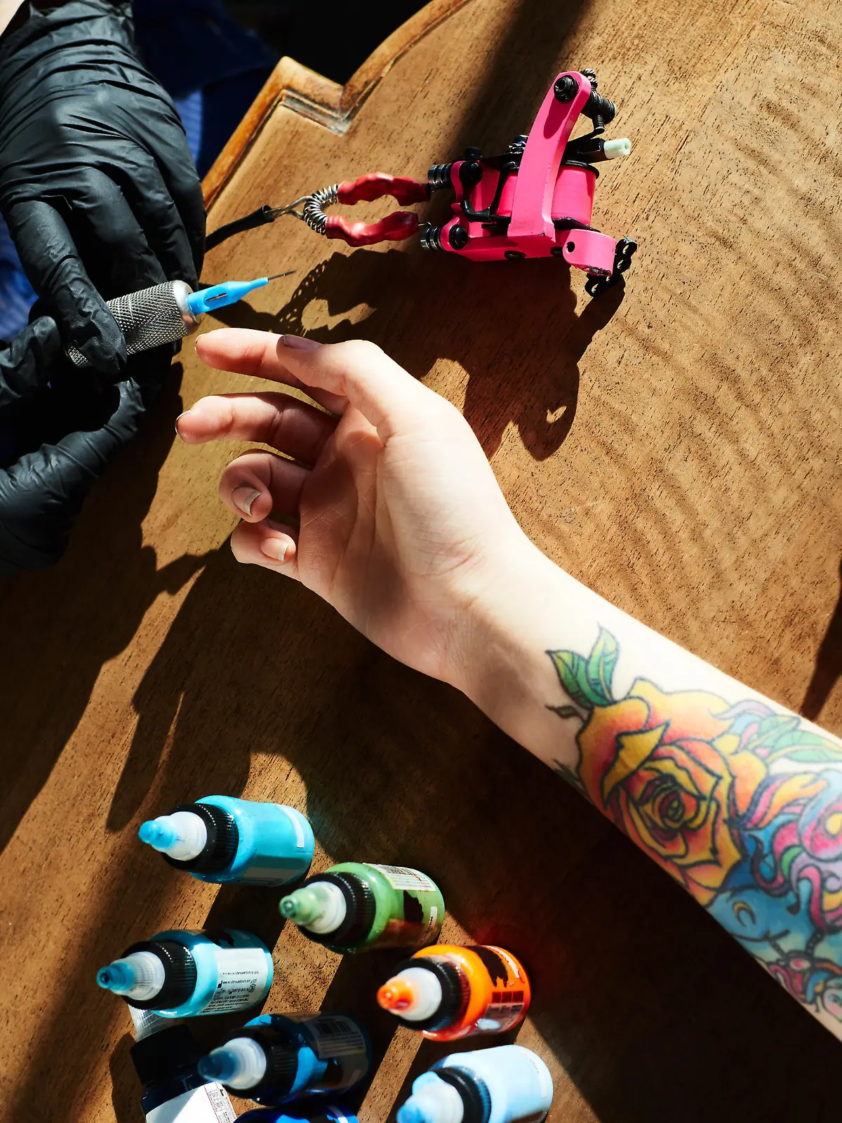 Tattoo expert in gloves holding tattoo gun while hand of female client with new colorful tattoo lying in wooden table beside, high angle close-up view