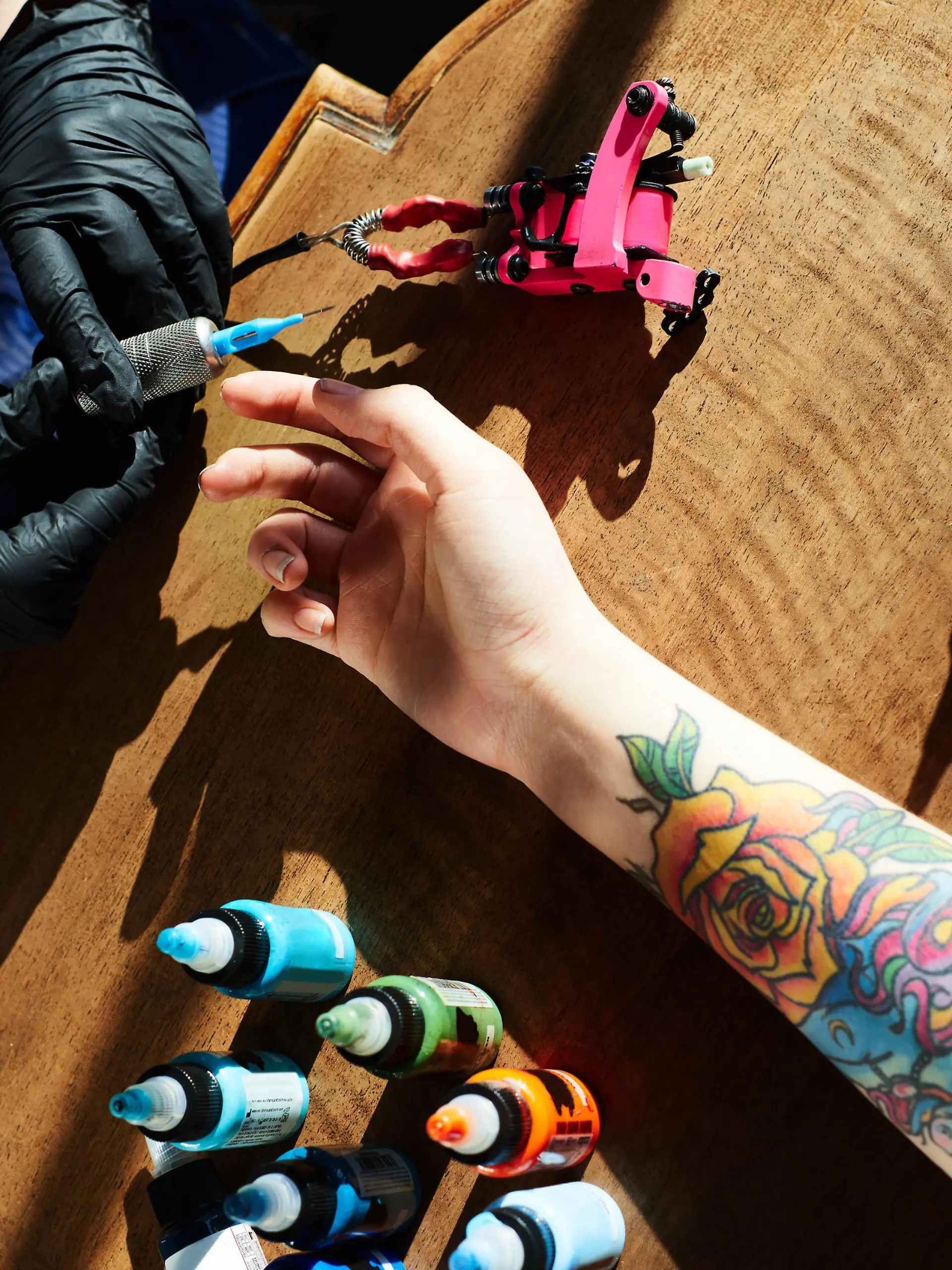Tattoo expert in gloves holding tattoo gun while hand of female client with new colorful tattoo lying in wooden table beside, high angle close-up view