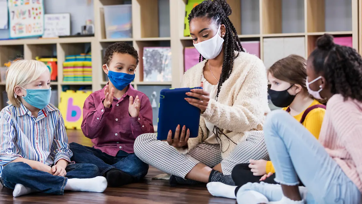 A multi-ethnic group of kindergarten or first grade students and their teacher in the classroom, sitting together in a circle on the floor. The children are 5 to 7 years old. They are all wearing masks, back to school during the COVID-19 pandemic, trying to prevent the spread of coronavirus. The teacher, an African-American woman, is holding a digital tablet and a boy is looking at it while clapping his hands.