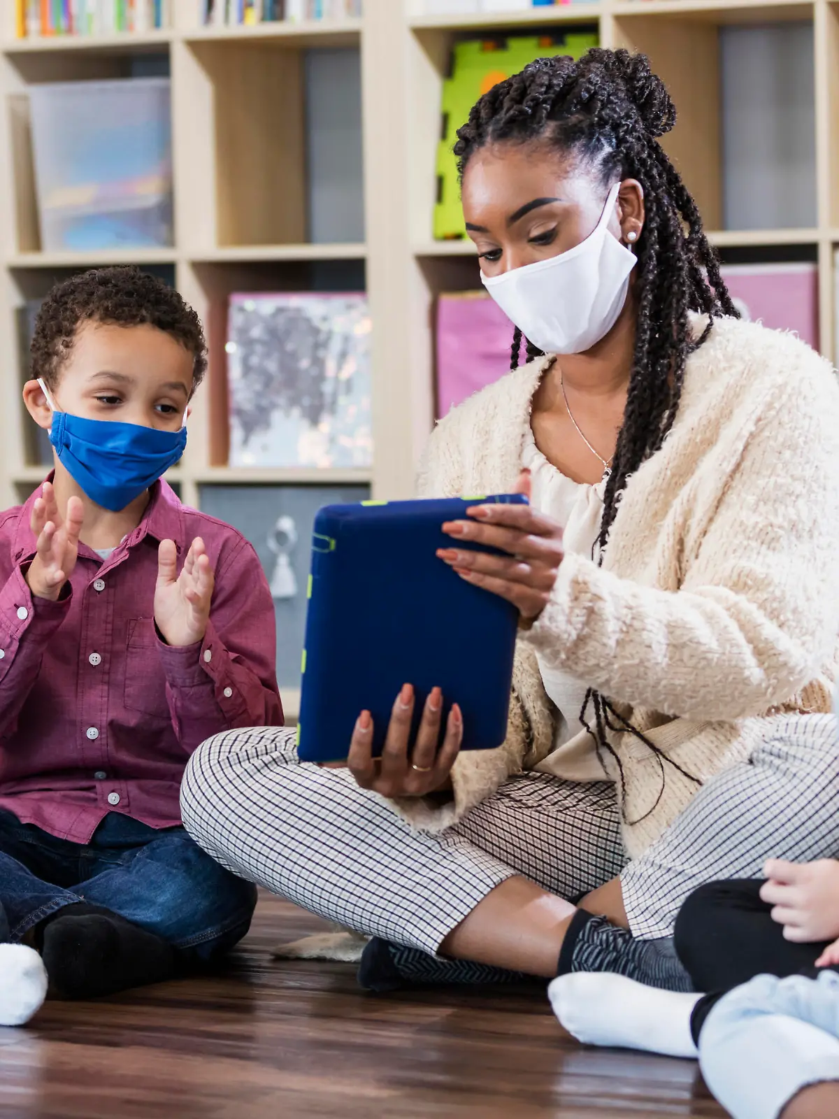A multi-ethnic group of kindergarten or first grade students and their teacher in the classroom, sitting together in a circle on the floor. The children are 5 to 7 years old. They are all wearing masks, back to school during the COVID-19 pandemic, trying to prevent the spread of coronavirus. The teacher, an African-American woman, is holding a digital tablet and a boy is looking at it while clapping his hands.
