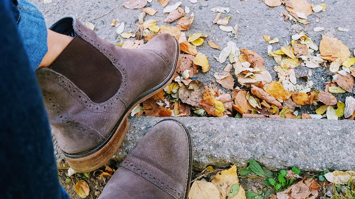 Ankle boots chelsea on legs in jeans on a background of asphalt and yellow autumn leaves.