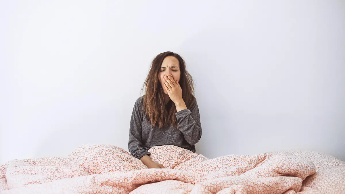 The girl is in bed and yawns. She covers her mouth with her hand. Early morning and she is sleepy.