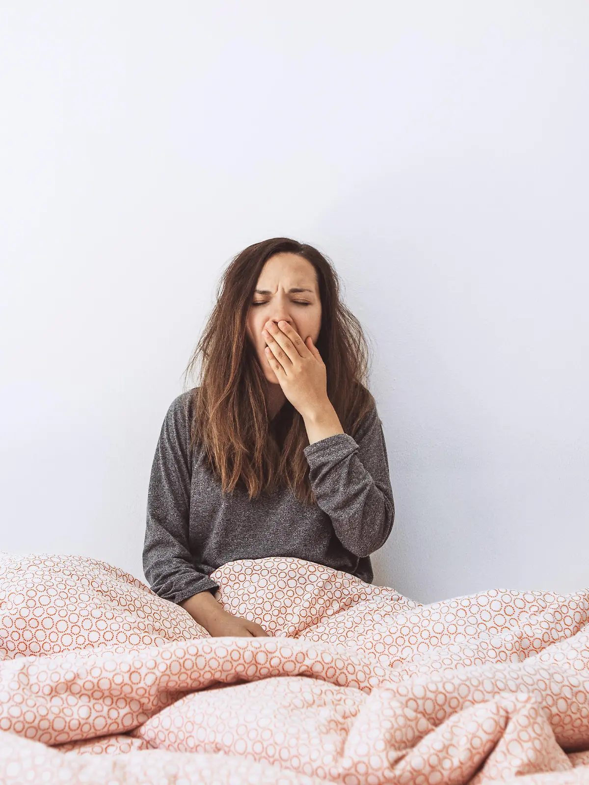 The girl is in bed and yawns. She covers her mouth with her hand. Early morning and she is sleepy.