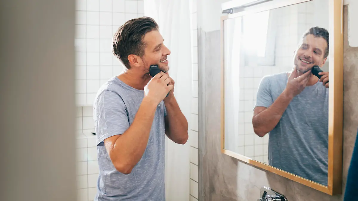 Handsome happy young Caucasian man shaving his beard with an electric razor in front of a bathroom mirror early in the morning.