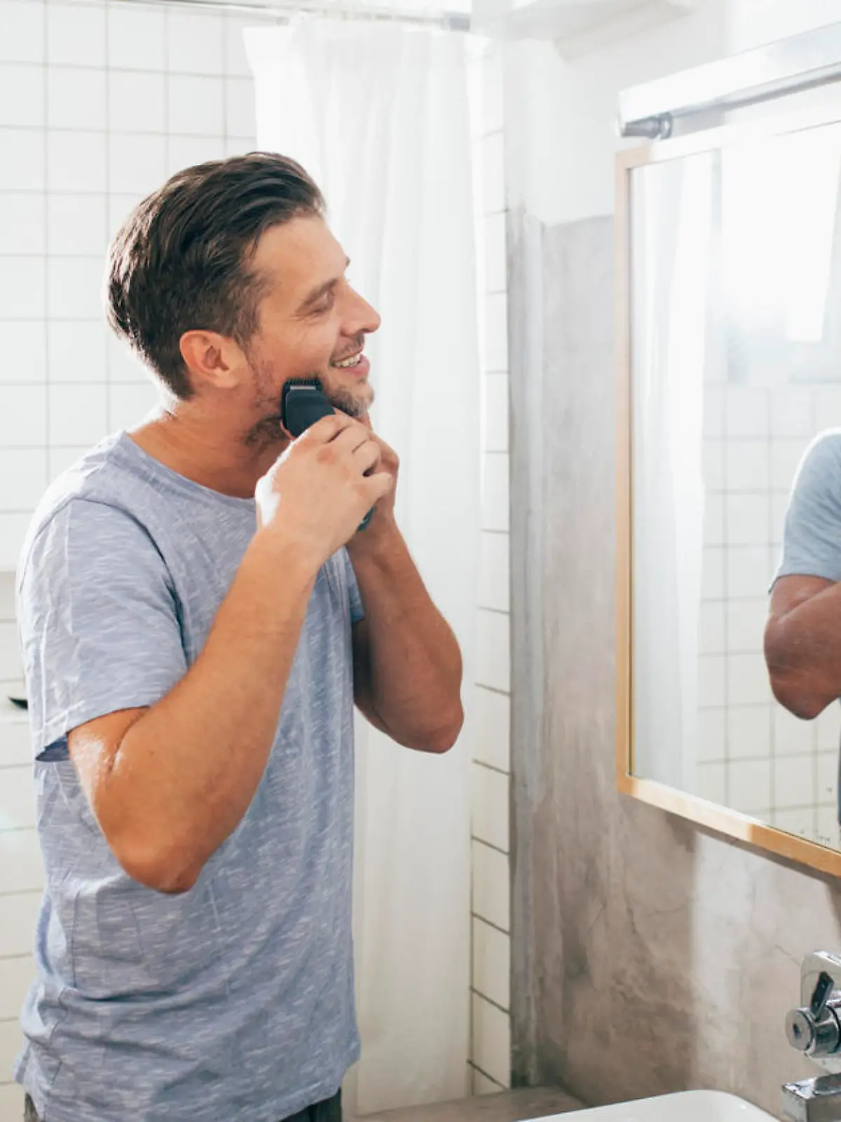 Handsome happy young Caucasian man shaving his beard with an electric razor in front of a bathroom mirror early in the morning.