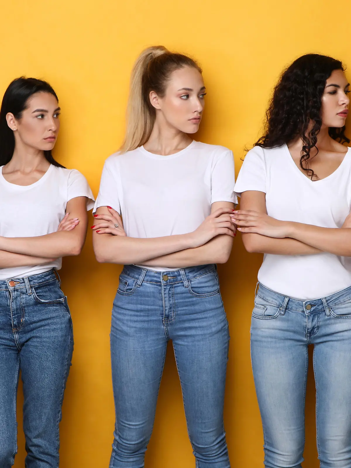 Offended Discontented Multiracial Ladies Looking At Asian Female Friend Unfriendly Disapproving Her Posing Crossing Hands Standing Over Yellow Studio Background. Adult Bullying And Racism