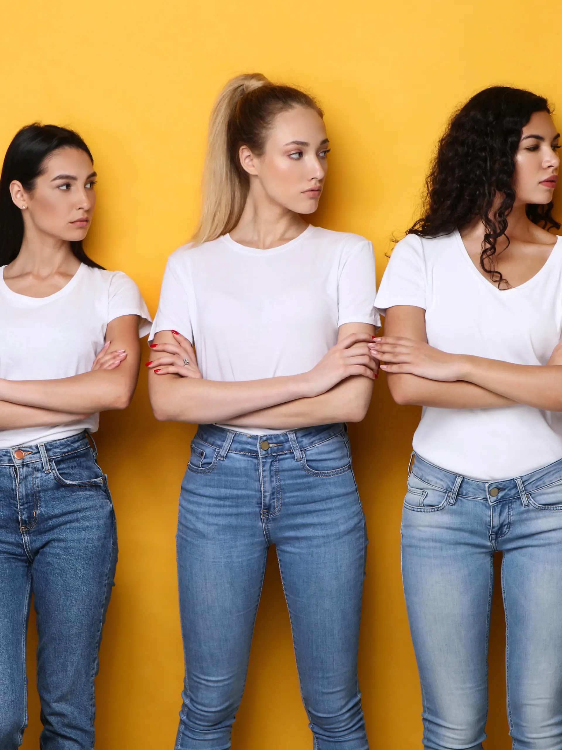Offended Discontented Multiracial Ladies Looking At Asian Female Friend Unfriendly Disapproving Her Posing Crossing Hands Standing Over Yellow Studio Background. Adult Bullying And Racism