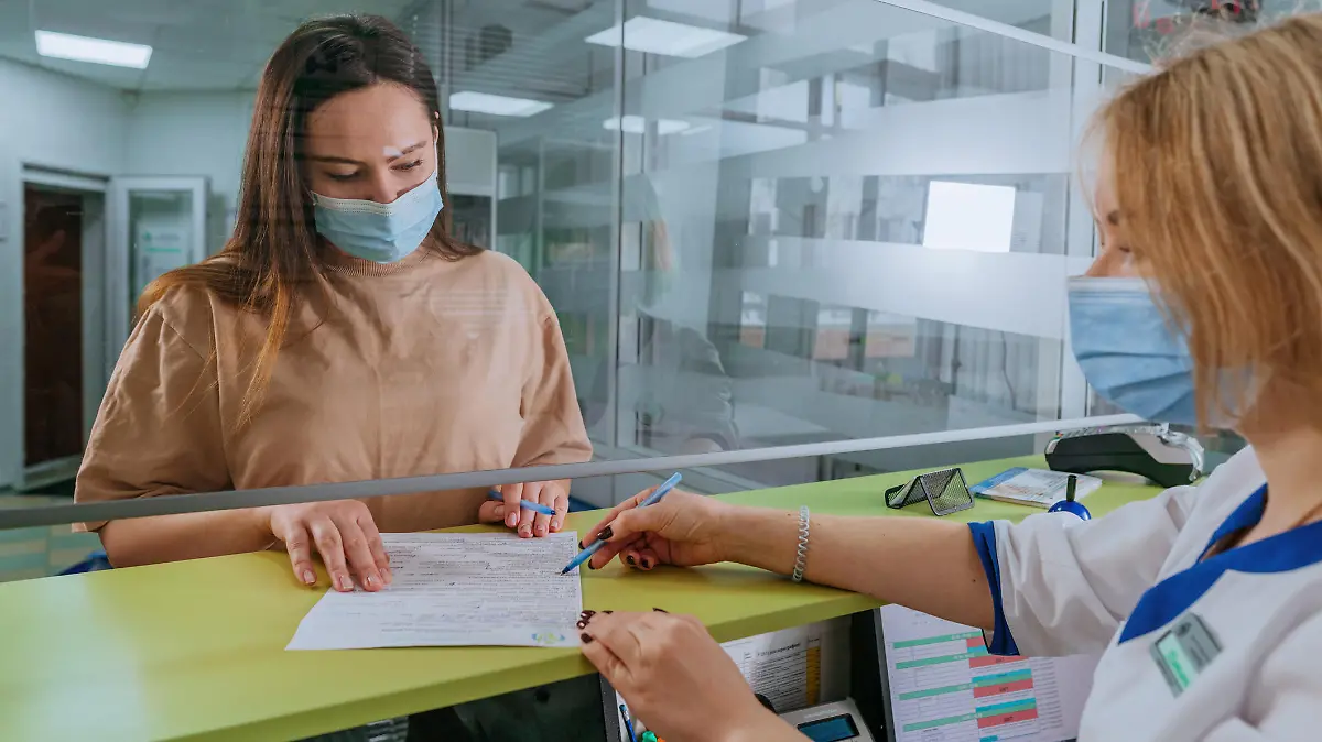 Female doctor or nurse explaining patient how to fill medical form at hospital reception. Doctor and patient wearing protective face masks and using protective glass screen. New normal during Pandemic COVID-19 concept. Selective focus on the patient.