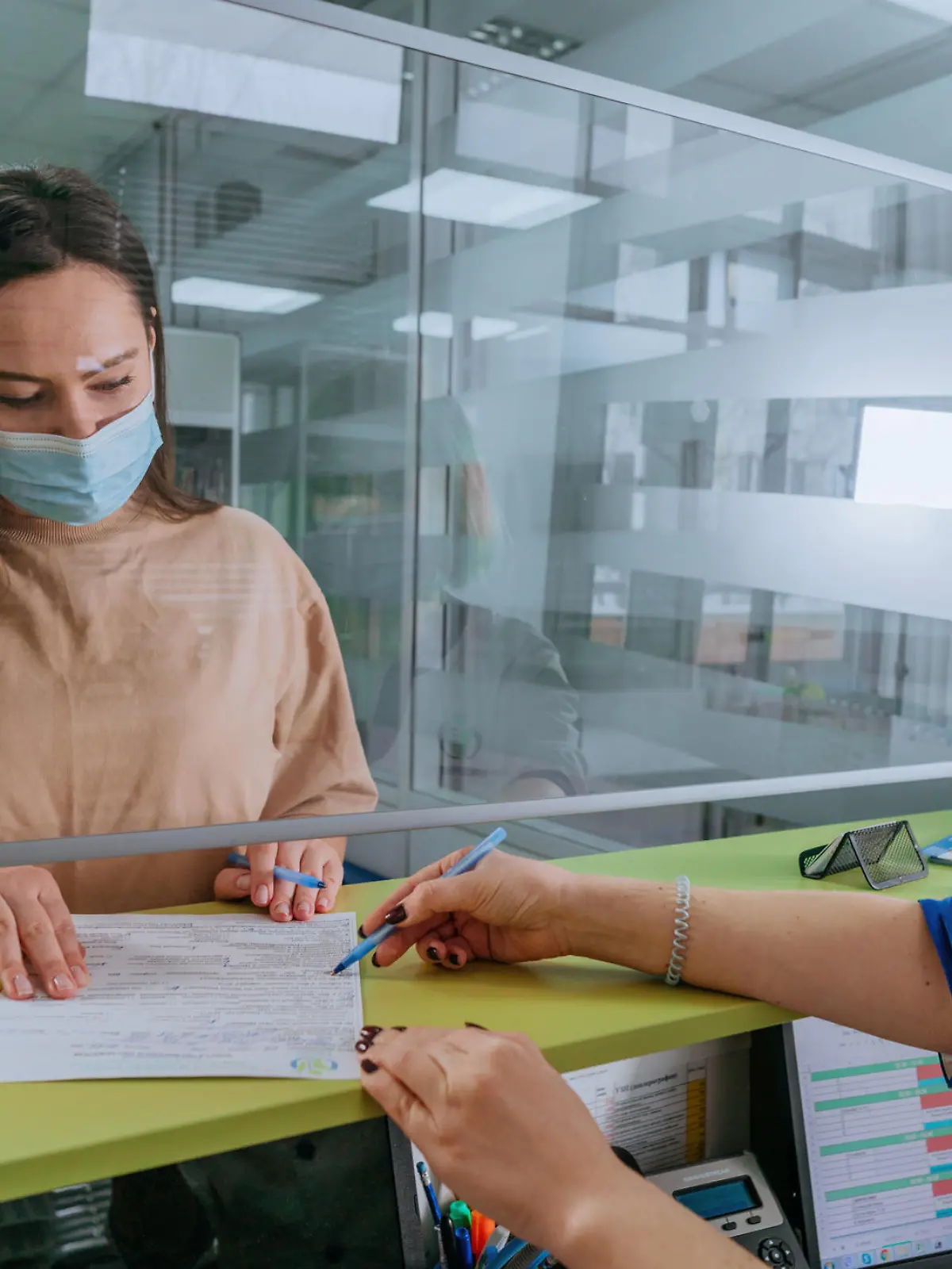 Female doctor or nurse explaining patient how to fill medical form at hospital reception. Doctor and patient wearing protective face masks and using protective glass screen. New normal during Pandemic COVID-19 concept. Selective focus on the patient.